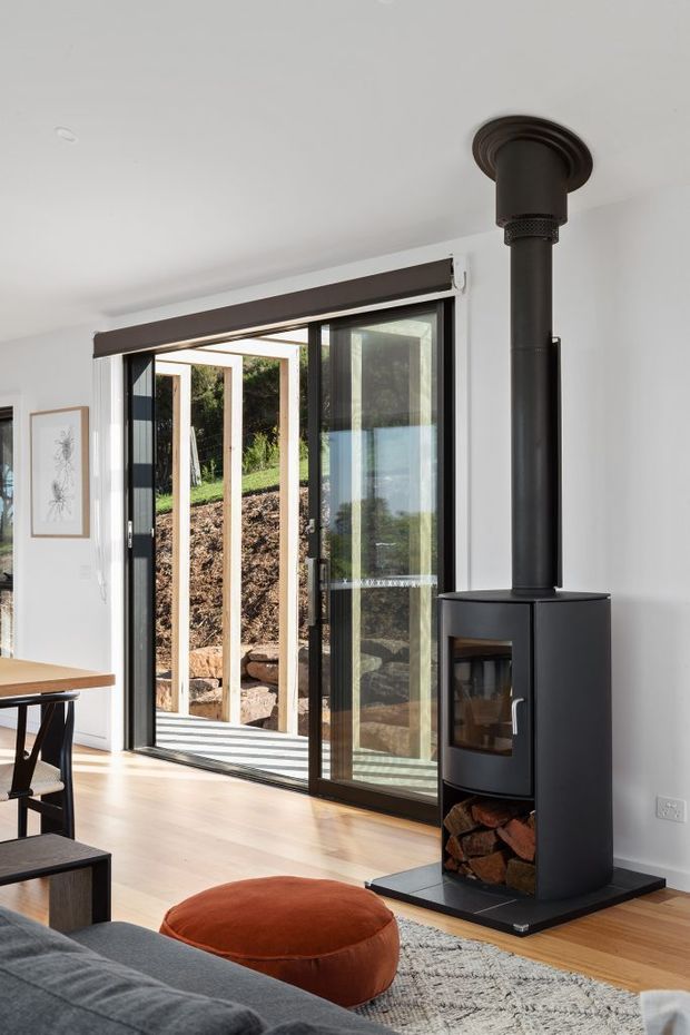 Bright contemporary living area with a freestanding black wood-burning stove and flue positioned beside full-height glazed sliding doors. The room has white walls, light timber flooring, a dark sofa, rust-coloured ottoman, rug, and a dining table partially visible at left. The glazing opens toward a landscaped outdoor area with exposed timber framing and stone retaining elements.