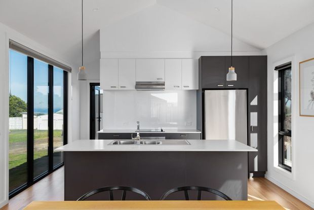 Bright contemporary kitchen with a large central island, white upper cabinetry, dark charcoal island and refrigerator surround, glossy white splashback, stainless-steel appliances, pendant lights, and extensive black-framed glazing opening toward a green outdoor setting.