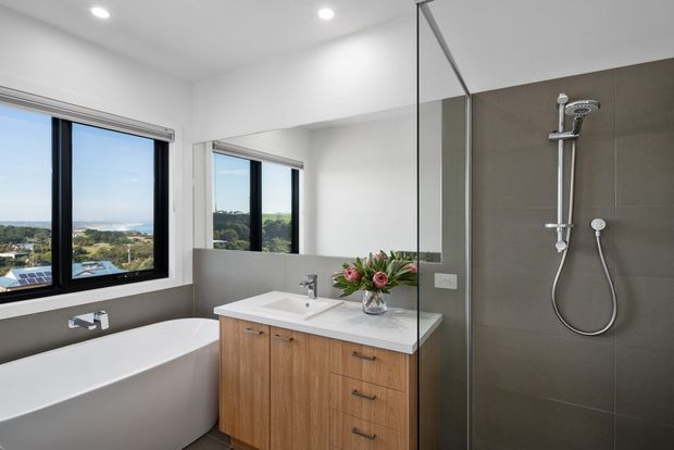 Bright contemporary bathroom with a freestanding white bathtub beside large black-framed windows, a wall-mounted shower within a frameless glass enclosure, and a timber vanity with a white benchtop. Large mirrors reflect the leafy suburban outlook and abundant natural light.