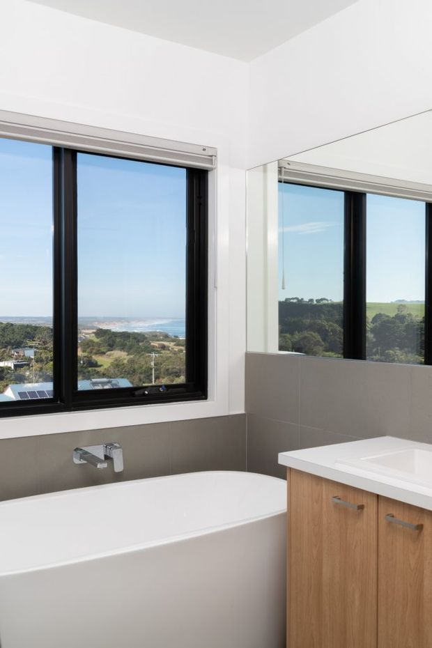 Bright contemporary bathroom with a built-in white bathtub beneath large black-framed windows, a timber vanity with a white countertop, grey wall tiles, chrome tapware, and a large wall mirror. The windows overlook a coastal landscape and ocean.