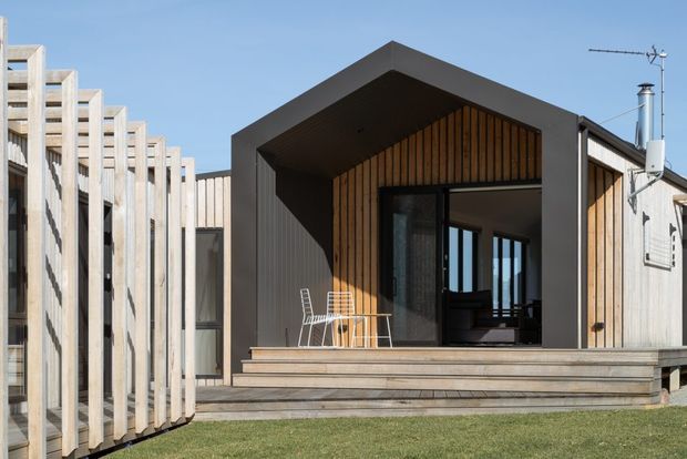 Exterior view of a compact contemporary timber-clad building with a bold dark gabled entry volume. The facade features vertical timber screening, a glazed recessed doorway, broad timber steps, and a small outdoor seating area.