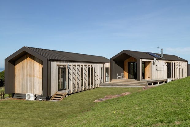 Exterior view of a low-profile contemporary residential building composed of connected gabled volumes on a gently sloping grassy site. The facade combines dark metal cladding with vertical light timber screening and timber-lined recessed entries. A raised timber deck, steps, large glazed openings, rooftop solar panels, and simple pitched roofs are visible.