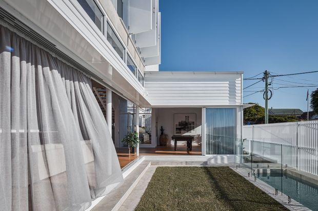 Exterior view of a contemporary multi-level home with strong horizontal lines, white cladding, extensive glazing, and a covered timber deck. Sheer curtains open onto the deck and an interior sitting or living area is visible through the glass. A narrow rectangular swimming pool with glass balustrading runs alongside a small lawn and paved walkway.