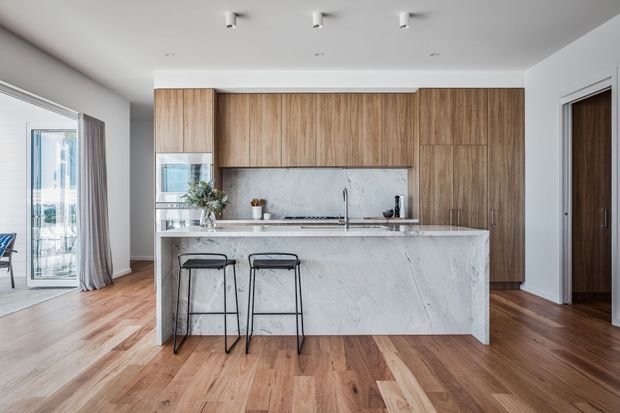 A bright, contemporary kitchen with a large white marble-look island, integrated sink and cooktop, two black bar stools, and full-height warm wood cabinetry. The kitchen opens toward a glazed outdoor area on the left, with timber flooring, white walls, recessed ceiling spotlights, and a dark timber interior door.
