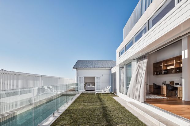 Bright exterior view of a contemporary multi-level residence with a rooftop or elevated terrace, glass balustrades, white horizontal cladding, and extensive sliding glass doors. The open living area visible through the glazing includes timber flooring, built-in timber joinery, a desk, and seating. A narrow grass terrace and a long linear water feature or pool run alongside the building, with a smaller gabled white-clad structure beyond.