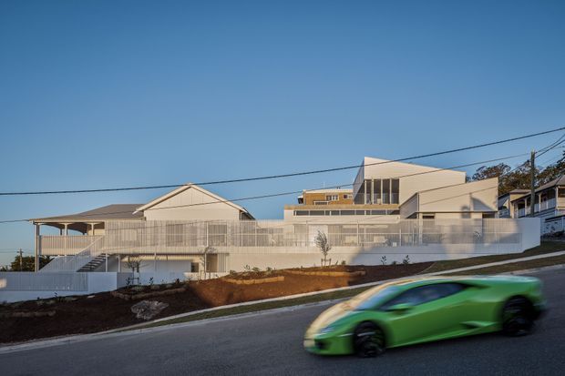Exterior street-level view of a large contemporary residential building on a sloped site. The composition shows multiple connected volumes, pale rendered walls, broad balconies and terraces with fine metal balustrades, large glazed openings, and a low-pitched roof form. A blurred green sports car passes in the foreground; overhead utility wires and neighbouring buildings are visible.