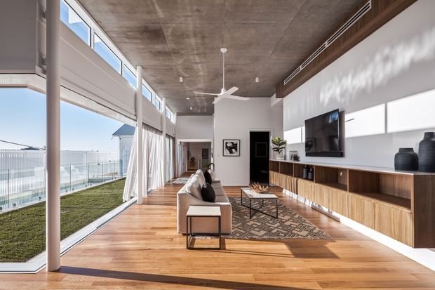 Bright contemporary living room with floor-to-ceiling glazing along one side, a timber floor, exposed concrete ceiling, white walls, and extensive built-in timber media and storage cabinetry. The room includes a sofa, coffee table, wall-mounted television, decorative shelving, and a ceiling fan. Glazing opens to a narrow outdoor strip and provides abundant natural light.