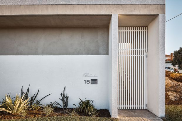 Street-facing entrance facade of a minimalist residential property, featuring a white rendered wall, warm grey exposed concrete canopy and upper band, and a tall white vertical slatted security or entry door. A small tiled threshold and drought-tolerant planting complete the restrained front entry composition.