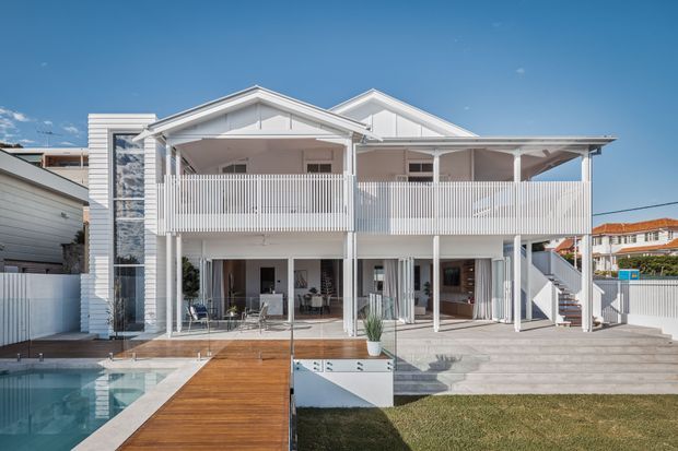 Exterior rear view of a large white multi-storey coastal-style residence with multiple pitched gables, broad covered verandas, white vertical balustrades, extensive glazing, and an external staircase. A swimming pool, timber deck and paved entertaining area occupy the foreground.