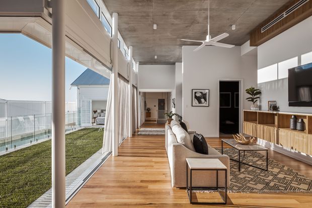 A bright contemporary living room with a long open-plan layout, extensive floor-to-ceiling glazing, timber flooring, a concrete ceiling, and a substantial built-in timber media cabinet. A sofa faces the television and coffee table, while a glazed outdoor area and lawn are visible beyond the windows.