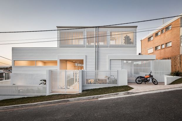 Street-facing view of a contemporary multi-level residence with a pale grey, horizontally clad facade and extensive vertical metal screening. The home features upper-level screened balconies or windows, a recessed illuminated entry, a gated front boundary, an enclosed garage or carport area, and a sloping suburban streetscape. A neighbouring exposed-brick apartment building is visible at right.