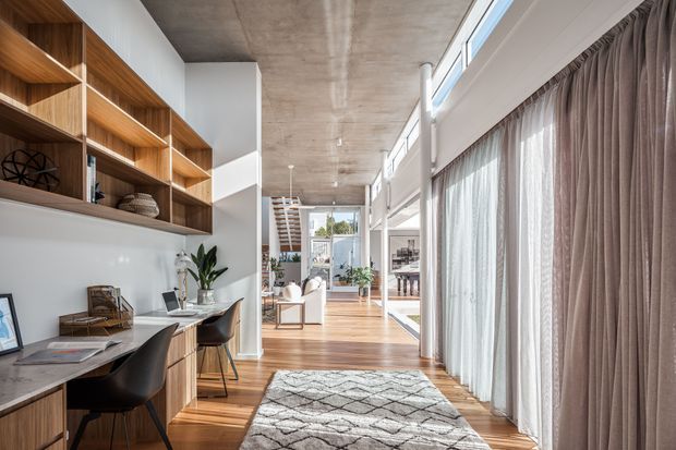 A bright, contemporary open-plan interior with a built-in timber study nook and shelving along the left wall, opening toward a living and dining area. Warm timber flooring, a textured concrete ceiling, expansive floor-to-ceiling glazing with sheer and taupe drapery, and a patterned rug create a refined, light-filled space. A staircase and further furnishings are visible in the background.