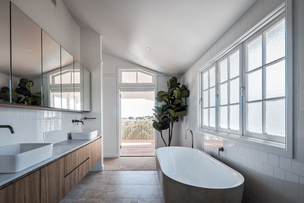 Bright contemporary bathroom with a freestanding oval bathtub positioned beneath a bank of white-framed frosted windows. The room features a double-basin timber vanity with mirrored cabinetry, white subway-tile wall finishes, pale stone-look flooring, dark tapware, and a glazed door opening to a small balcony or deck. A tall indoor plant adds a natural accent.
