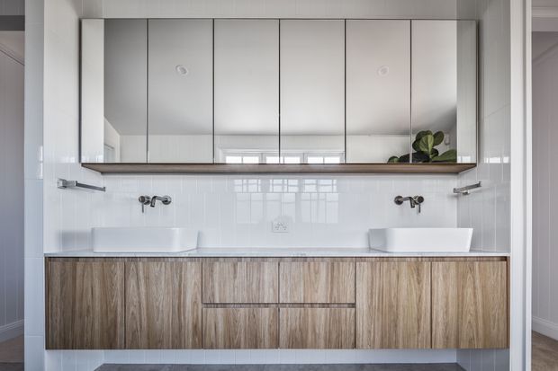 A bright contemporary double-sink bathroom vanity with extensive mirrored cabinetry above a long timber-look built-in vanity. White vessel basins, wall-mounted chrome tapware, glossy white rectangular wall tiles, and a dark horizontal shelf create a clean, symmetrical composition. A doorway and hallway are visible at the sides, with a plant reflected in the mirror.