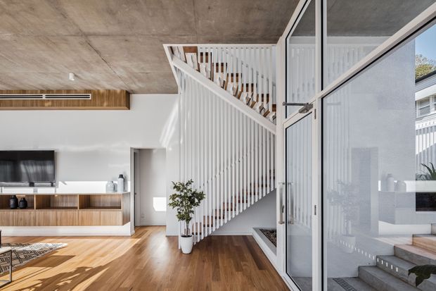Bright contemporary open-plan living area with warm timber flooring and built-in timber media cabinetry, a white TV wall, and a prominent white slatted staircase with open balustrade. A large glazed pivot or hinged door opens toward an exterior entry or courtyard, while a raw concrete ceiling and crisp white walls create a clean, modern interior. Natural daylight fills the space.