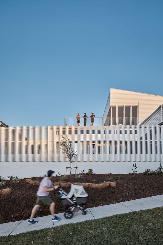 Exterior architectural photograph of a contemporary multi-level residential building with pale horizontal cladding, extensive white vertical slatted screening and a rooftop terrace with three people. A landscaped frontage, footpath and person pushing a stroller appear in the foreground beneath a clear blue sky.