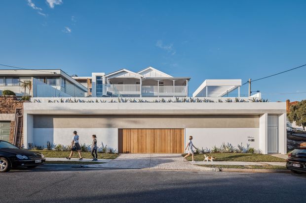 Street-facing view of a contemporary multi-level residential building with a broad flat-roofed garage or lower-level facade, smooth light rendered walls, a large warm timber garage door, narrow vertical window opening, and planted roof-edge landscaping. Upper levels behind it feature pitched gabled roof forms, balconies with pale metal or timber balustrades, and extensive glazing. The image is photographed from across a paved street in clear daylight.