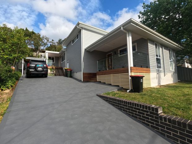 Exterior view of a contemporary multi-level residential home on a sloping suburban site. The house has light grey horizontal weatherboard cladding, a shallow pitched roof with broad eaves, a covered front porch with timber-toned screening, and a dark paved driveway bordered by a low black brick retaining wall. Mature landscaping and a parked vehicle are visible.