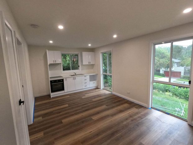 A bright, unfurnished open-plan room with a compact white kitchen along the back wall, integrated oven and cooktop, white cabinetry, and a grey benchtop. Warm brown wood-look flooring runs throughout, with recessed ceiling lights and large windows and glazed doors overlooking greenery and neighbouring homes.