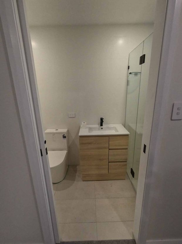 Compact contemporary bathroom viewed through its doorway, featuring a white toilet, floating light-wood vanity with white integrated basin and black tapware, large pale floor tiles, and a frameless glass shower enclosure with dark hardware.
