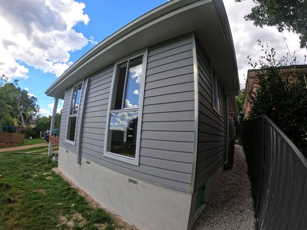 Exterior view of a compact single-storey house or extension with grey horizontal weatherboard cladding, white-trimmed vertical windows, a low-slope curved roof edge, and a concrete base. A narrow gravel side passage and timber boundary fencing are visible alongside the building.