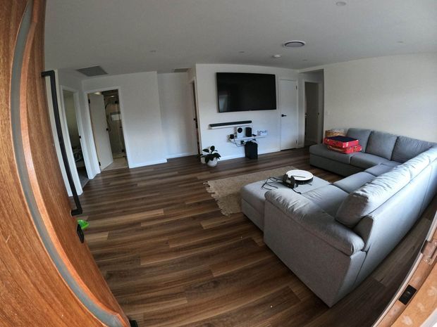 A bright contemporary living room viewed through a timber-framed doorway, featuring dark wood-look flooring, white walls and ceiling, a large grey sectional sofa, wall-mounted television, low media console, and a small rug. Doorways lead to adjacent rooms in the background.
