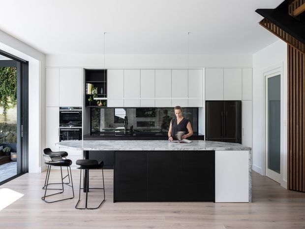 Bright contemporary kitchen with a large central island, white full-height cabinetry, black lower joinery, a dark reflective splashback, and a pale stone island benchtop. Two black bar stools sit at the island, with integrated appliances and a full-height black refrigerator along the rear wall. Pale timber flooring, pendant lights, and a timber slatted feature wall add warmth and texture. Glazed doors and windows bring in abundant natural light.
