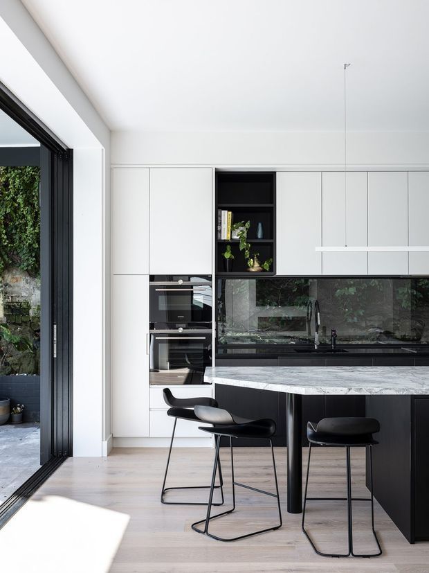 Bright contemporary kitchen with white full-height cabinetry, integrated black appliances, a black-framed open glass door, dark reflective splashback, pale timber flooring, and a large light marble-look island with black cabinetry below. Three black bar stools sit at the island, while greenery and a stone garden wall are visible outside.