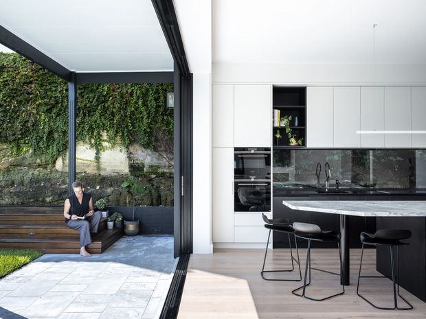 Bright contemporary kitchen opening directly onto a covered outdoor courtyard through fully retractable black-framed glazing. The interior features white full-height cabinetry, integrated black appliances, a dark stone-look splashback and benchtop, a large island with waterfall edge, and black bar stools. Pale timber flooring continues toward the threshold, while the outdoor area has light paving, a built-in dark bench, planted greenery, and a stone retaining wall.
