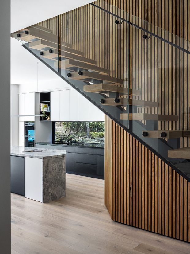 A contemporary open-plan kitchen viewed beneath a striking floating staircase. The staircase has thick timber treads, dark metal framing and clear glass balustrades, set against a full-height vertical timber-slat feature wall. The kitchen includes white and dark-grey flat-panel cabinetry, a large veined stone island, a black-framed window, and pale timber flooring. The composition highlights the stair structure and timber wall while showing the kitchen beyond.