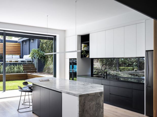 Contemporary kitchen with a large marble island and waterfall end, dark lower cabinetry, white overhead cabinets, and a reflective dark splashback. A broad glazed opening connects the kitchen to a landscaped outdoor courtyard, with two bar stools at the island. The space is bright, refined, and strongly connected to the garden.