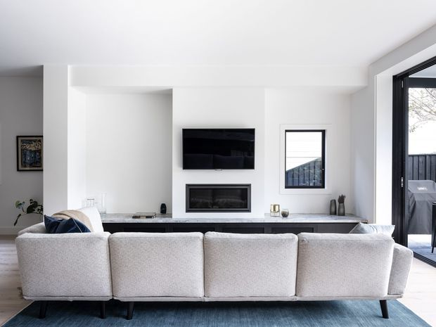 A bright, contemporary living room viewed from behind a large light-grey sectional sofa. The room features white walls and ceiling, a minimalist built-in entertainment wall with a wall-mounted television and recessed fireplace, a long pale stone-look ledge, dark-framed windows and glazed doors, and light timber flooring. A blue-grey rug and restrained decor add subtle contrast.