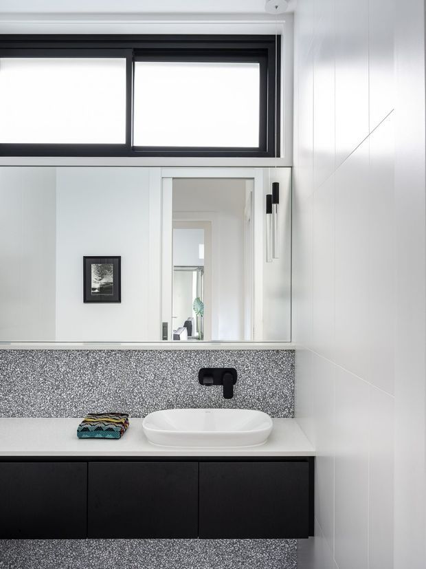A bright, contemporary bathroom vanity viewed straight on. The composition features a floating matte-black vanity with a white vessel basin, black wall-mounted tapware, a speckled grey mosaic splashback, white wall panelling, a large mirrored cabinet, and black-framed clerestory windows. A narrow view through the doorway reveals another bright interior space.