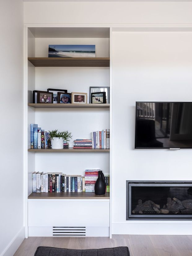 Bright, clean built-in living room wall featuring recessed open shelving with timber shelves, framed family photographs, books, and a small plant. Beside it is a wall-mounted flat-screen television above a contemporary linear fireplace with a dark surround. Pale painted walls and light timber flooring create a calm, minimal interior.