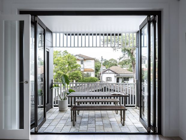 View from a bright interior through fully opened black-framed glazed doors onto a tiled terrace or balcony. The outdoor space has a grey metal balustrade, a long picnic-style dining table with benches, and a potted plant. Neighbouring homes and trees are visible beyond the railing. The interior has white walls and a light timber floor, creating a clean indoor-outdoor connection.