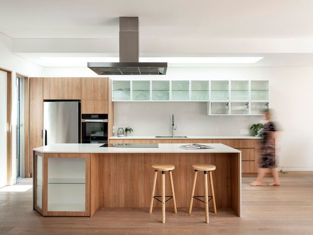 Bright contemporary kitchen with a large timber-clad island, white stone benchtop, integrated stainless-steel appliances, open glass-fronted upper cabinetry, and timber flooring. The kitchen is photographed from the living area, with a skylight or linear ceiling light and floor-to-ceiling glazing at the side.