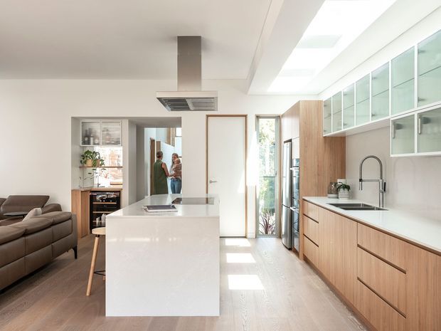Bright, contemporary open-plan kitchen viewed from the main cooking area, with a large white island, integrated cooktop and ceiling-mounted rangehood, extensive light timber cabinetry, pale benchtops, glass-fronted overhead cupboards, stainless-steel appliances, and a full-height glazed side wall with skylights. A living area and people are visible beyond the kitchen, with warm timber flooring throughout.