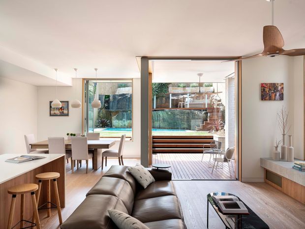 Bright open-plan contemporary interior combining a kitchen, dining area and living room. The space features pale timber flooring, white walls, a timber kitchen island with stools, a dining table, a brown leather sectional sofa, pendant lights, built-in media joinery and a ceiling fan. Large glazed sliding doors open to a timber deck and a visible swimming pool, bringing substantial natural light into the room.