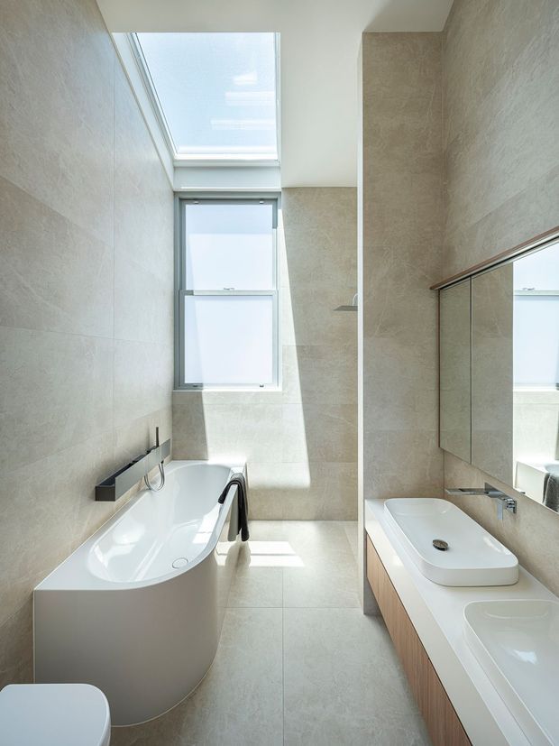 A bright, narrow contemporary bathroom featuring a freestanding white bathtub, dual white vessel basins on a timber-toned vanity, large wall-to-wall mirrors, pale stone-look surfaces, and a skylight with tall frosted windows. Black tapware and fittings provide contrast, while natural daylight creates a clean, airy atmosphere.