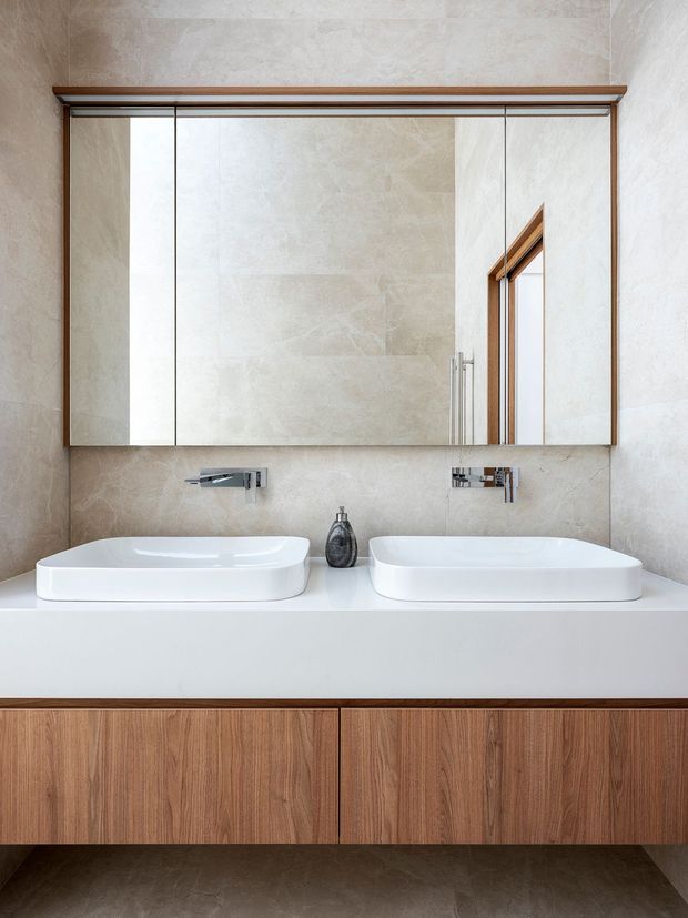 Contemporary bathroom vanity featuring two white vessel basins on a white countertop, wall-mounted chrome taps, a large mirrored cabinet, warm timber joinery, and pale stone-look wall and floor finishes. The composition is clean and symmetrical with soft neutral tones and natural wood accents.