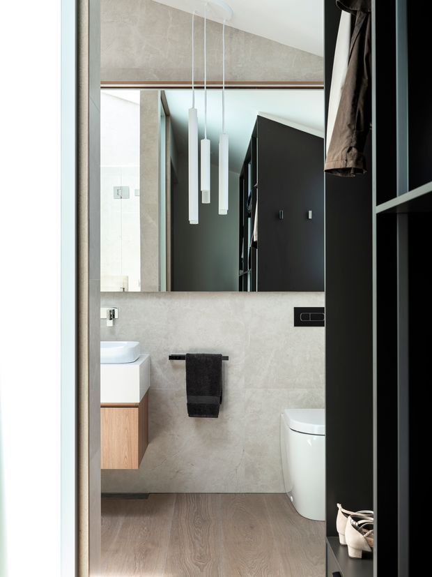 A contemporary bathroom viewed through a doorway, featuring pale stone-look wall surfaces, warm timber vanity cabinetry, a white basin and toilet, a large mirror, black shelving, and slim white pendant lights. The room has a refined monochrome palette with warm wood flooring and dark accents.