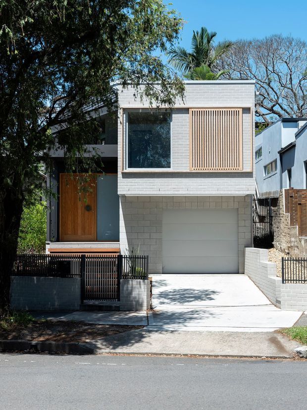 Street-facing view of a compact multi-storey urban residence with a light grey painted-brick facade, integrated garage, white concrete driveway, timber front door, and a large upper-level window partly screened by vertical timber battens. Black metal fencing, rendered side walls, and mature trees frame the contemporary townhouse-like frontage.