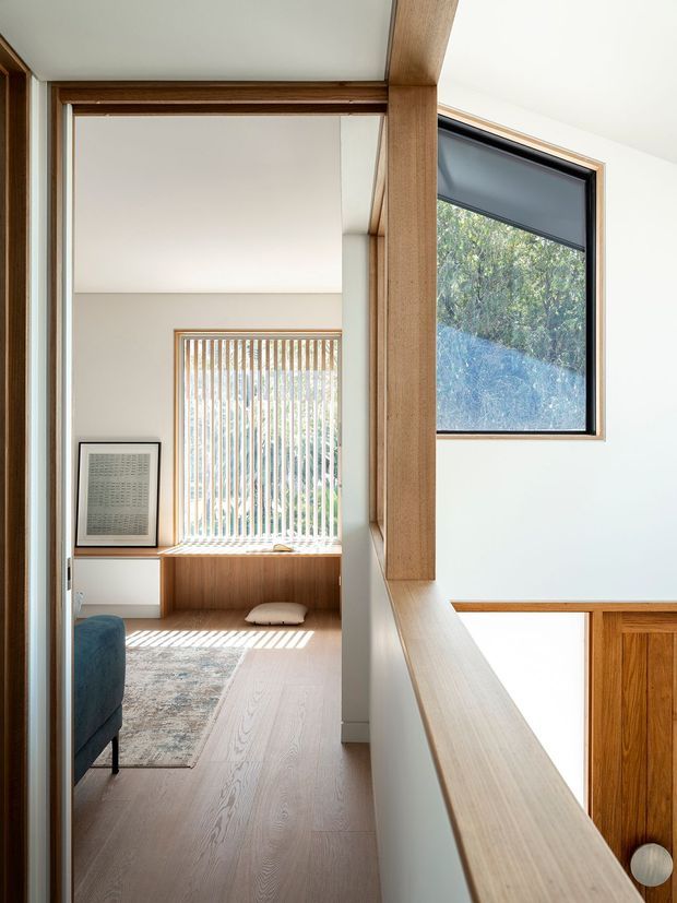 Bright interior view looking through a timber-framed opening toward a small sitting or living area. Pale timber flooring, warm timber framing and built-in window seating contrast with white walls and ceilings. Vertical timber screening filters daylight at the rear window, while a dark-framed side window overlooks greenery. A partial blue upholstered chair, patterned rug, framed artwork and timber cabinetry are visible.