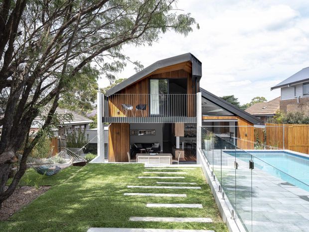 Exterior view of a contemporary two-storey timber-clad residence with a sharply sloping roof, upper balcony, open lower-level living area, landscaped lawn with stepping stones, and an adjacent swimming pool enclosed by clear glass fencing. The facade combines warm vertical timber, dark metal roofing, and light structural elements.
