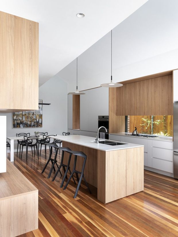 Bright contemporary kitchen with a large timber island, white benchtops, integrated timber cabinetry, stainless-steel appliances, and a dining area with black chairs. The kitchen opens into a high-ceilinged open-plan space with warm timber flooring, pendant lights, and a window above the rear bench.