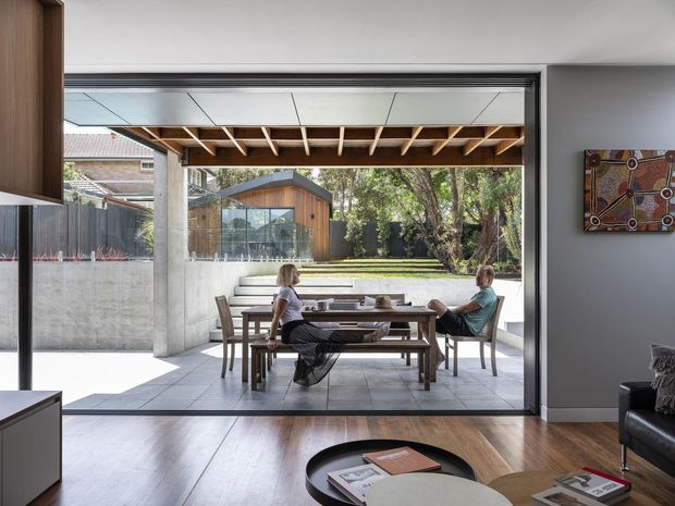 A contemporary indoor-outdoor living and dining space with broad retractable glazing opening onto a paved courtyard. The foreground has dark timber flooring, a low round coffee table and partial seating, while the covered outdoor area contains a timber dining table and chairs. Exposed timber roof framing, concrete columns and large sliding glass panels create a strong connection to the landscaped garden beyond.