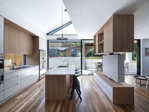 Bright contemporary open-plan kitchen and living space with a central island, white waterfall benchtop, extensive timber cabinetry, integrated appliances, timber flooring, and a large glazed opening to an outdoor area. A sculptural angled ceiling and high-level glazing bring in abundant natural light.