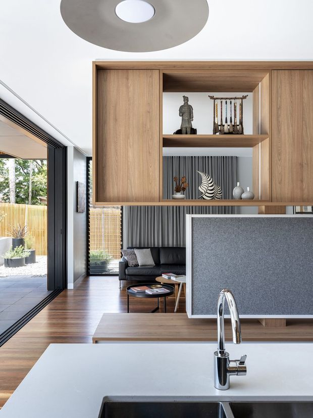 A contemporary open-plan kitchen viewed from the island, with a white benchtop and chrome mixer tap in the foreground. Warm timber flooring and built-in timber cabinetry with open display shelving frame views toward a dark sofa and living area. A grey textured partition separates the kitchen, while full-height black-framed glazing opens to an outdoor patio. The room has a large circular ceiling light and a restrained palette of timber, white, grey and black.
