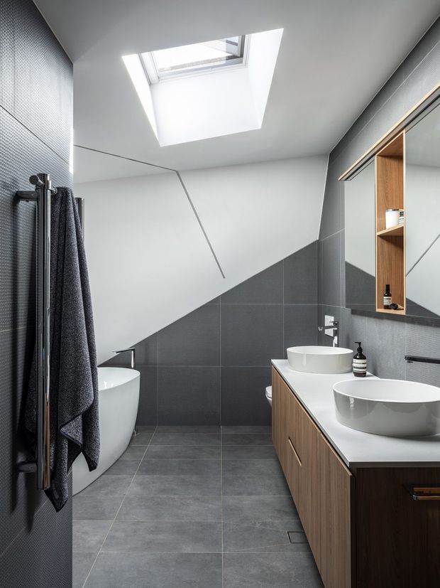 Contemporary bathroom with a sloped ceiling and large skylight, featuring dark grey tiled walls and floor, a freestanding white bathtub, double white vessel basins on a timber vanity, mirrored storage, and a vertical towel warmer. The room has a clean, refined monochromatic palette with warm wood joinery accents.