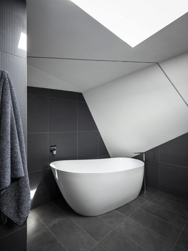 A contemporary bathroom with a freestanding white oval bathtub, dark charcoal wall and floor tiles, minimalist chrome tapware, and a dramatic angular white ceiling incorporating a skylight. A textured dark towel hangs at the left edge of the frame.